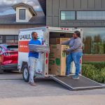 A couple begins unloading their cargo trailer in the driveway of their new home. The woman unloads a U-Haul TV moving box, and the man carries some folded furniture blankets.