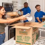 Two Service Providers help a customer pack her plates and glasses into U-Haul moving boxes in the kitchen. The customer puts towels into one of her boxes. Packing tips for moving include keeping your plates standing and never packing them flat.