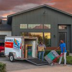 A man and a woman unload their belongings from their U-Haul trailer and into their new home. Is it better to rent or buy a house in 2024? It’ll depend on many factors.