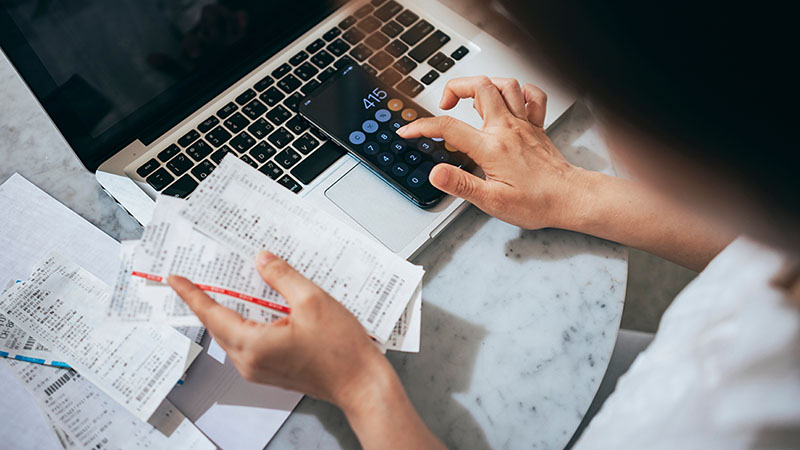 A woman uses the calculator on her phone to review her bills and receipts. Her phone sits on her computer, which sits on a table.