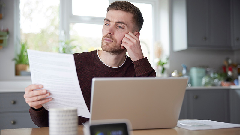 A man sits at a table while holding a paper bill with his laptop open in front of him on the table. The cost of living depends on several factors, including housing, food, health care, and transportation.