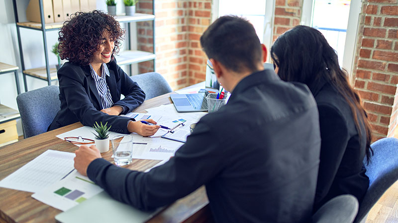 A real estate agent explains the documents in front of a couple while using a pen to point at the documents. Housing mortgage rates will remain between 6 to 7 percent in 2024.