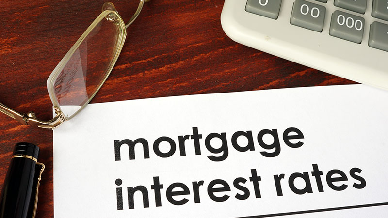 The words “mortgage interest rates” are written on a piece of paper that sits on a desk. Surrounding the piece of paper is a calculator, a pair of glasses, and a pen.”