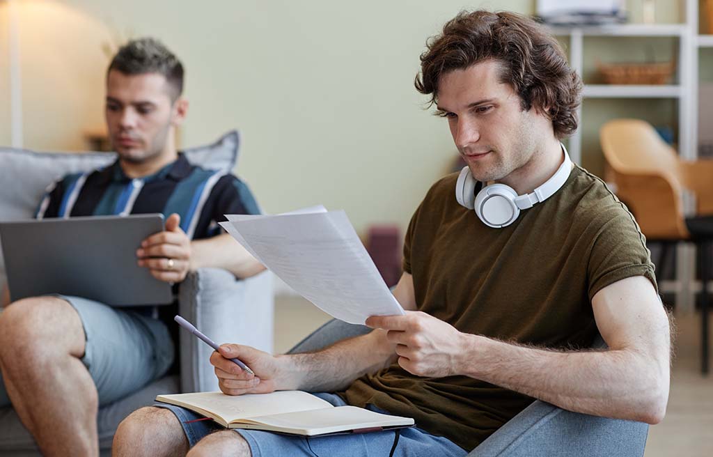 Two roommates study together in the living room. One roommate reviews his paper notes while his notebook sits on his lap. The other roommate reviews his homework on his computer.