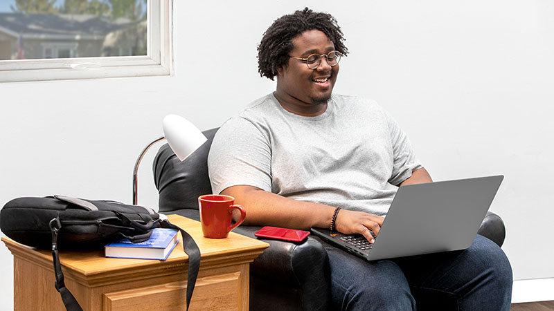 A male freshman sits down in a chair while typing on his computer. A desk lamp sits on an end table, which is one of many dorm room essentials that Moving Help recommends purchasing.