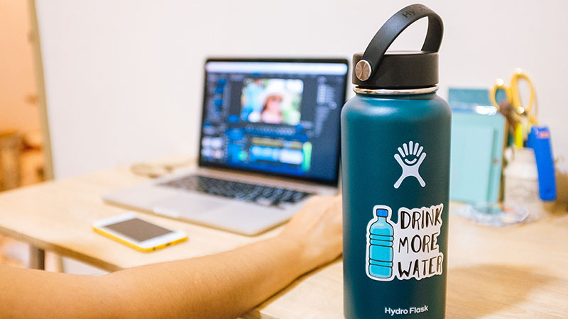 A Hydro Flask water bottle sits on a desk as a college student works on the computer. A portable travel cup is one of the dorm room essentials Moving Help recommends purchasing.