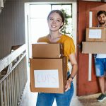 A woman and a man carry moving boxes labeled cups and books as they move into their new urban home.