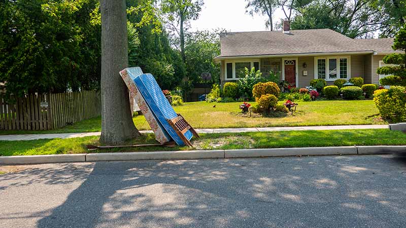 Two old mattresses and part of a bed frame sit on the curb by a house. In some areas, curbside pick-up from the city is available to help you get rid of your mattress.