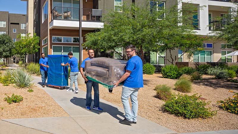 Two furniture Moving Helpers carefully move a couch outside and to a customer’s U-Haul truck rental. In the background, two moving labor providers also are carefully moving a piece of furniture on a furniture dolly.