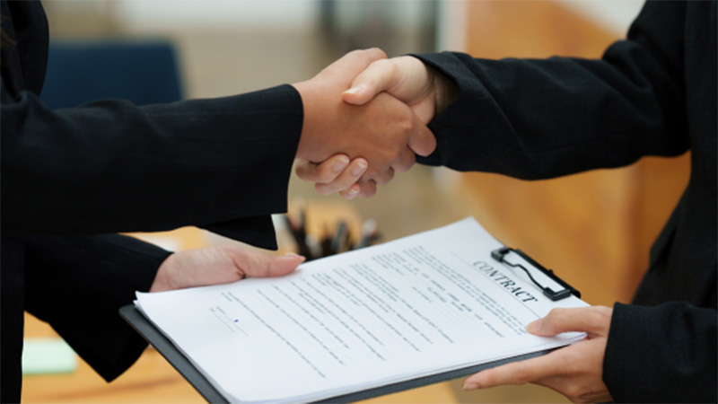 Two people shake hands while holding onto a clipboard with signed paperwork.