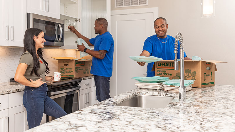 Two Moving Helpers work together to pack glasses and dishes in the kitchen of a customer’s home. The customer smiles while giving her helpers packing instructions for her kitchen.